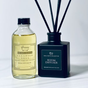 May include: A black glass room diffuser bottle with black reeds and a white label that reads "SOUL BATH COLLECTIVE ROOM DIFFUSER SOULBATHCOLLECTIVE.COM". Next to it is a clear glass bottle with a black cap and a white label that reads "SOUL BATH COLLECTIVE CLEANSED SOUL BERGAMOT + CLARY SAGE + EUCALYPTUS ROOM DIFFUSER OIL SOULBATHCOLLECTIVE.COM 1 fl oz".