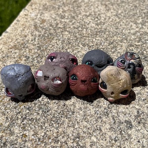 May include: A collection of seven small, sculpted alien heads in various colors, including gray, brown, and red. Each head has large, expressive eyes and unique surface textures, creating a whimsical and otherworldly aesthetic.