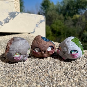 May include: Three small, hand-painted ceramic figurines with whimsical faces. Each figurine is shaped like a rock with different colors and textures. One figurine is brown with blue and white eyes, another is gray with pink and white eyes, and the last is gray with green and white eyes.