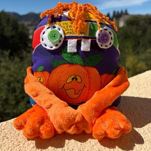 May include: A plush toy with a purple fabric body featuring a pumpkin print, orange textured arms, and orange yarn hair. It has two white felt eye patches with a green button and a purple button, plus two white felt teeth and text that reads SIN Y ELFRY URN UNGLON.