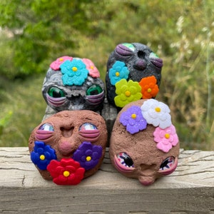 May include: Four small, hand-painted, ceramic figures with whimsical faces and colorful flower accents. Each figure has a unique design and expression.