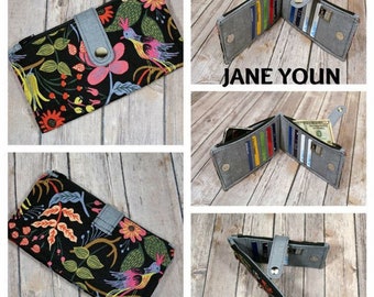 May include: A black fabric wallet with a floral and bird print. The wallet has a zipper closure, a snap closure, and a gray fabric flap with a snap closure. The wallet is open to show the interior, which has multiple card slots and a cash compartment. The wallet is on a wooden surface. The text "JANE YOUN" is visible on the image.