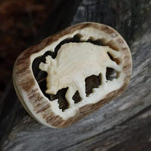 May include: A carved antler pendant featuring a detailed relief of a bison. The bison is cream-colored, set against a dark background within a brown antler frame. The pendant is displayed on a weathered wooden surface.
