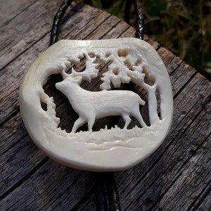 May include: A white bone bolo tie featuring a carved deer in a forest scene.