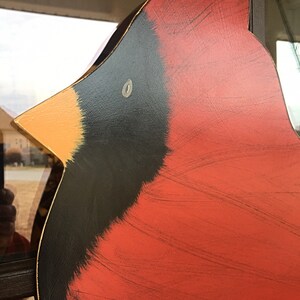 May include: Close-up of a red cardinal bird wooden cutout with black and yellow accents. The bird is painted with a distressed look and has a white eye. The text "Birds Nest Door Decor" is visible at the bottom of the image.