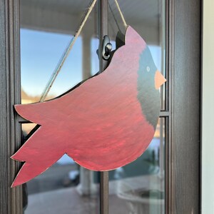 May include: A red cardinal bird wooden cutout with black accents hanging on a door with a brown frame.