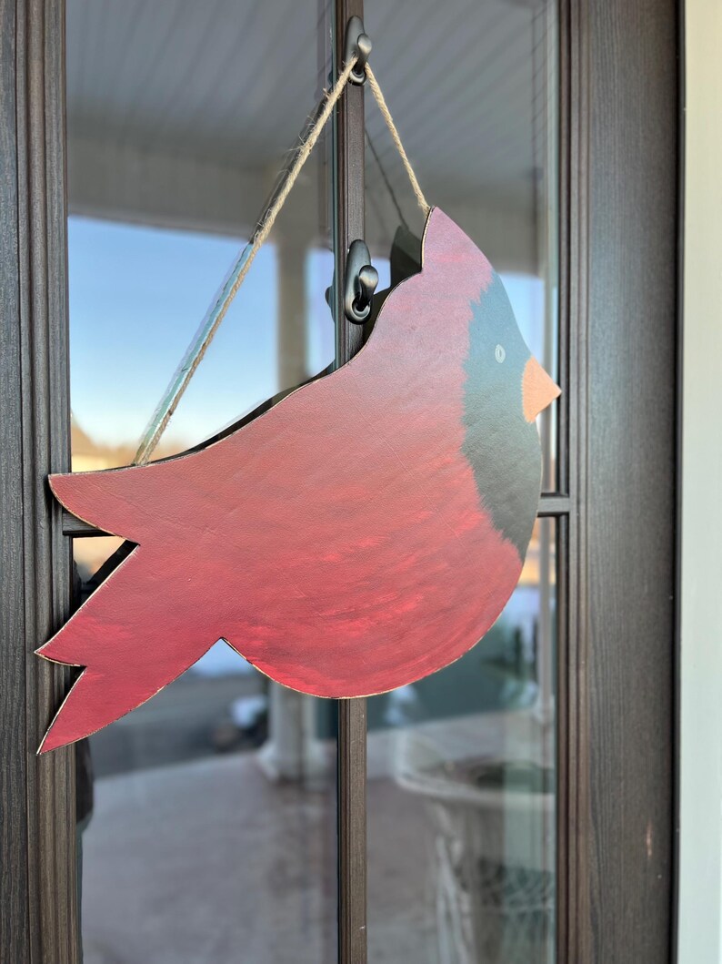 May include: A red cardinal bird wooden cutout with black accents hanging on a door with a brown frame.