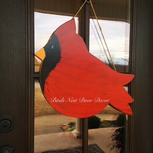 May include: A red cardinal bird wooden door hanger with black and yellow accents. The hanger is hanging from a string and has the text "Birds Nest Door Decor" written on it.