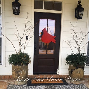 May include: A red cardinal wooden door hanger with a black beak and yellow accents hangs on a brown front door with a glass panel. The door is flanked by two potted plants with green leaves and brown pots. The door is surrounded by white siding and there is a brown doormat with the text "Birds Nest Door Decor" on it.