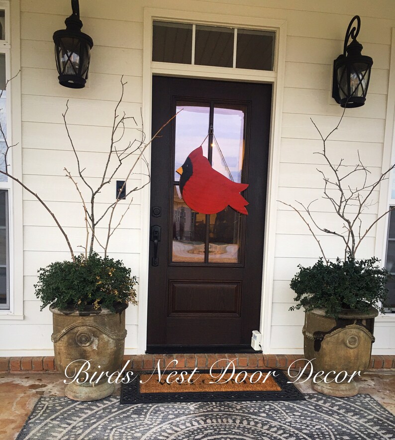 May include: A red cardinal wooden door hanger with a black beak and yellow accents hangs on a brown front door with a glass panel. The door is flanked by two potted plants with green leaves and brown pots. The door is surrounded by white siding and there is a brown doormat with the text "Birds Nest Door Decor" on it.
