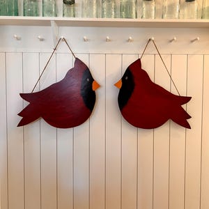 May include: Two red wooden cardinal bird ornaments with black eyes and yellow beaks hanging on a white wall with white vertical panels.