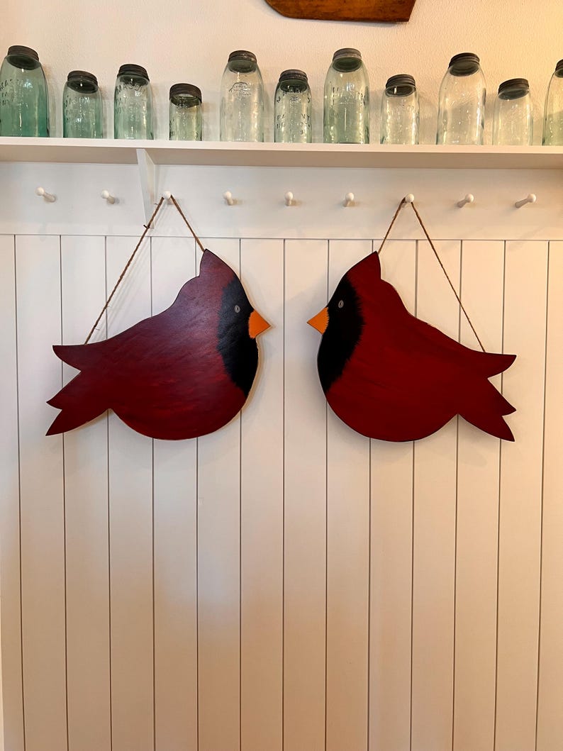 May include: Two red wooden cardinal bird ornaments with black eyes and yellow beaks hanging on a white wall with white vertical panels.