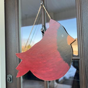 May include: A red cardinal bird cutout with black accents hanging on a door. The bird is made of wood and has a rustic, hand-painted look.