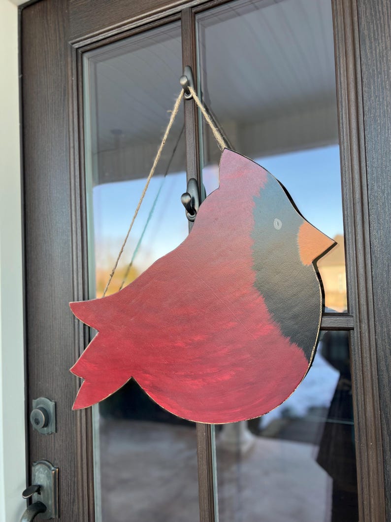 May include: A red cardinal bird cutout with black accents hanging on a door. The bird is made of wood and has a rustic, hand-painted look.