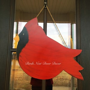 May include: Red cardinal bird wooden door hanger with black eyes and beak. The hanger is painted with a distressed look and has the text "Birds Nest Door Decor" written on it.