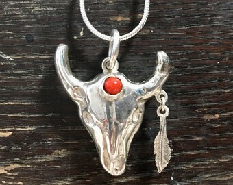 Handmade silver cow skull shaped pendant inset with red stone and wearing a silver feather earring