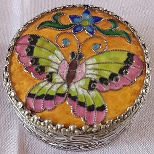 May include: A circular trinket box with a silver filigree base and an enamelled lid. The lid displays a butterfly design with pink, green, and black wings on a yellow background, with a blue flower accent. A decorative item.
