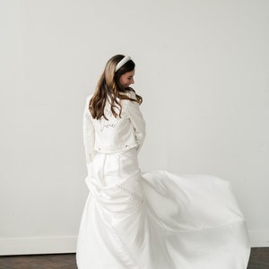 Pode incluir: Uma noiva com um vestido de noiva branco e uma jaqueta jeans branca combinando com o texto "Mrs. Dovie" nas costas. Ela est&aacute; usando uma faixa de cabe&ccedil;a branca e est&aacute; sorrindo. O vestido tem uma longa cauda.