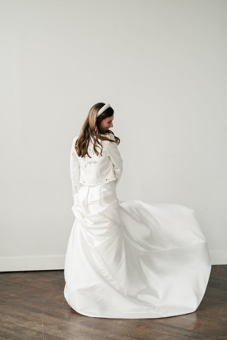 Pode incluir: Uma noiva com um vestido de noiva branco e uma jaqueta jeans branca combinando com o texto "Mrs. Dovie" nas costas. Ela est&aacute; usando uma faixa de cabe&ccedil;a branca e est&aacute; sorrindo. O vestido tem uma longa cauda.
