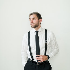 May include: A man wearing a white dress shirt with a black tie and black suspenders. The suspenders have the initials "JMD" on them. He is wearing black pants and a black belt.