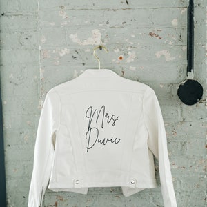 Pode incluir: Jaqueta jeans branca com o texto "Mrs. Duvic" em escrita preta nas costas. A jaqueta est&aacute; pendurada em um cabide dourado contra uma parede de tijolos desgastada. Possui fecho de z&iacute;per e detalhes de bot&otilde;es.
