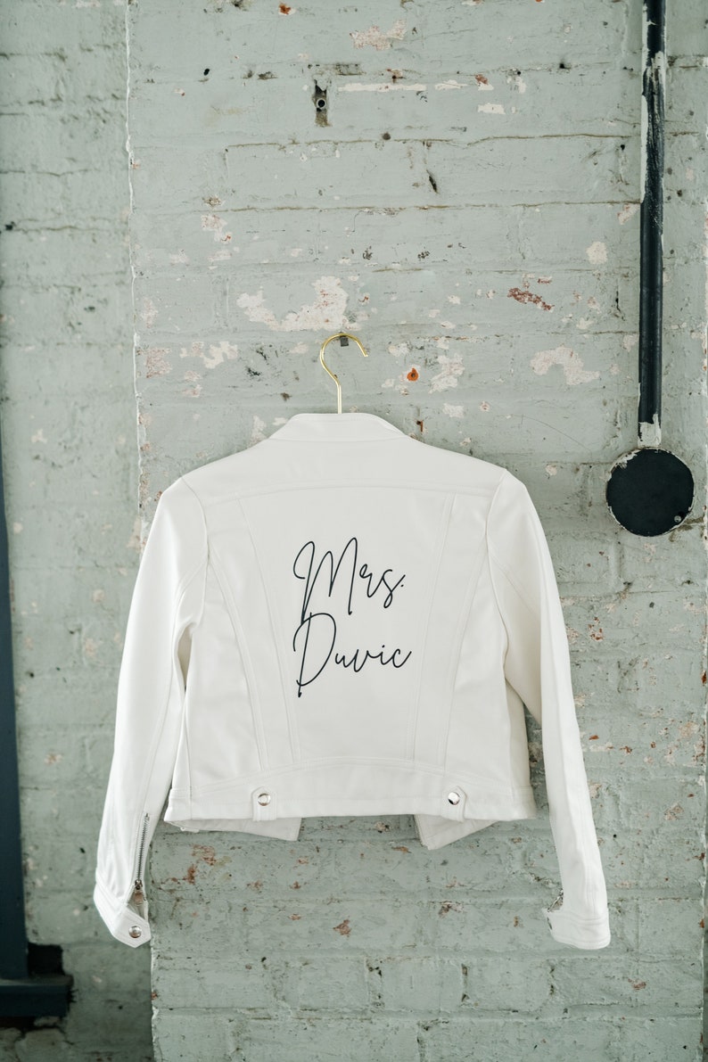 Pode incluir: Jaqueta jeans branca com o texto "Mrs. Duvic" em escrita preta nas costas. A jaqueta est&aacute; pendurada em um cabide dourado contra uma parede de tijolos desgastada. Possui fecho de z&iacute;per e detalhes de bot&otilde;es.