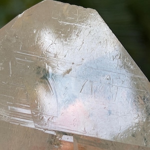 Può includere: Una punta di cristallo di quarzo trasparente e traslucido con una terminazione angolare affilata. La superficie presenta intricati motivi geometrici incisi, striature orizzontali e sfaccettature naturali che catturano la luce, creando una texture riflettente.