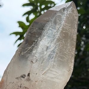 May include: A large, elongated clear quartz crystal point with a translucent, multifaceted surface. The crystal features natural internal inclusions, visible fractures, and a pointed termination, displaying a mix of clear and smoky grey tones.