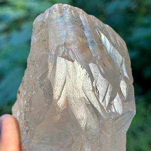 Può includere: Un grande cristallo di quarzo traslucido con una texture naturale ruvida. Il cristallo è tenuto in mano, mostrando la sua forma irregolare e la struttura cristallina interna. Il colore è un bianco pallido e chiaro con sottili variazioni.