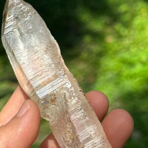 May include: A clear quartz crystal point with a natural, raw finish. The crystal is long and slender, with a pointed tip and a flat base. It has a light brown colour and is translucent.