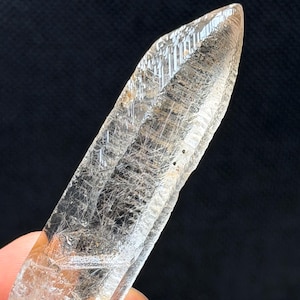 May include: A clear, elongated quartz crystal point featuring visible internal needle-like inclusions and distinct geometric surface striations. The crystal has a pointed termination and a translucent appearance, showcasing natural mineral formations.