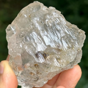 May include: A clear, rough-cut quartz crystal with a layered, textured appearance. The crystal is held in a hand, showcasing its natural, irregular shape and translucent quality. The background is a blurred green, suggesting an outdoor setting.