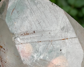 Puede incluir: Un cristal de cuarzo transparente con patrones e inclusiones internas visibles. El cristal tiene una punta afilada y una superficie rugosa y texturizada. El fondo es verde borroso, lo que sugiere un entorno natural al aire libre.