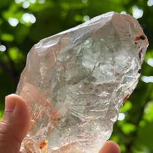 Puede incluir: Un cristal grande y transparente con una forma rugosa e irregular. El cristal es translúcido, permitiendo el paso de la luz. Tiene una apariencia pálida, casi incolora, con algunas áreas de tonos marrón claro y naranja. El cristal se sostiene en una mano sobre un fondo verde borroso.