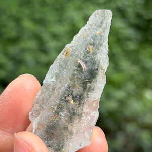 May include: A pale green, translucent crystal with an irregular shape. It features inclusions of darker minerals and tiny gold flecks. The edges are rough and uneven, giving it a natural, unpolished appearance. The crystal is held in a hand.