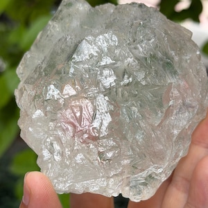 Può includere: Un cristallo di fluorite grezzo, non tagliato, dall'aspetto traslucido e vetroso. Il cristallo ha una tonalità verde pallido con alcune aree chiare. La superficie è ruvida e strutturata, con strutture cristalline visibili. Il cristallo è tenuto in una mano.