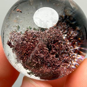 Pode incluir: Uma esfera de vidro ou cristal transparente contendo um aglomerado denso e texturizado de inclusões minerais castanho-avermelhadas profundas com aspeto metálico. O interior apresenta uma forma circular branca opaca suspensa.