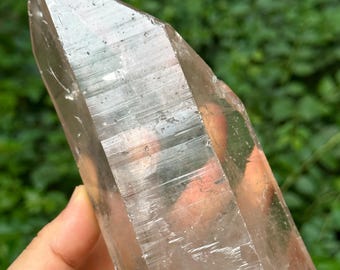 Puede incluir: Un gran cristal de cuarzo transparente con una punta. El cristal tiene una superficie facetada y un aspecto ligeramente ahumado. El fondo es un verde borroso, lo que sugiere un entorno natural al aire libre. El cristal se sostiene en una mano.