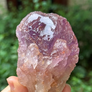 May include: A large, raw amethyst crystal with a purple and clear colour gradient. The crystal has a rough, natural texture and is partially translucent.