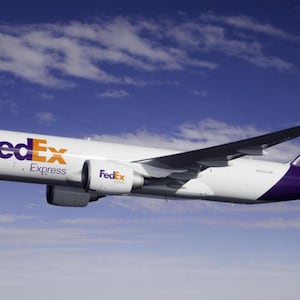 May include: A FedEx Express cargo airplane in flight against a blue sky with scattered clouds. The aircraft is white with purple and orange branding, including the FedEx logo on the fuselage and tail.
