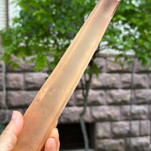 Może przedstawiać: Duży, brzoskwiniowy kryształ z ostrym zakończeniem. Kryształ trzymany jest w dłoni, ukazując swój wydłużony kształt i naturalną, nieoszlifowaną fakturę. Tło przedstawia zielone liście i ceglany mur.