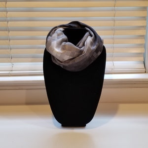 May include: A gray and white plaid infinity scarf displayed on a black mannequin. The scarf is made of a soft material and has a repeating pattern of squares. The scarf is draped in a circular shape.