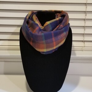 May include: A plaid infinity scarf in shades of purple, orange, yellow, and green. The scarf is displayed on a black mannequin head. The fabric has a soft, textured appearance, and the pattern is a classic plaid design.