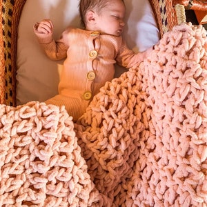 Può includere: Un bambino che dorme in una culla intrecciata, che indossa una tutina color pesca con bottoni di legno. Una coperta e un cuscino in maglia grossa abbinati, color pesca, circondano il bambino, creando un ambiente accogliente e confortevole.