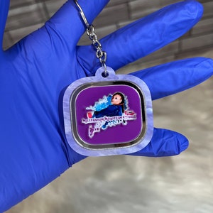 May include: A white and silver square keychain with a purple background featuring a person's face and the text "Millennium Manifestations".