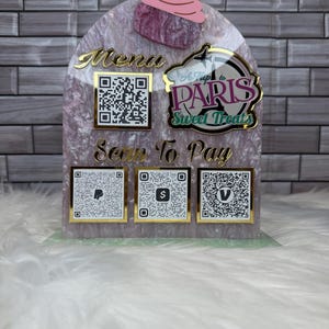 May include: A pink and white sign with the words "A Trip to Paris Sweet Treats" and a cupcake graphic. The sign also has a QR code labeled "Menu" and three QR codes labeled "Scan To Pay" for different payment methods.