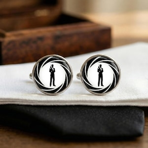 Peut inclure: Deux boutons de manchette argentés avec un motif noir et blanc. Chaque bouton de manchette présente une silhouette d'une personne tenant une arme, entourée d'un motif en spirale noir et blanc. Ils reposent sur une chemise blanche.