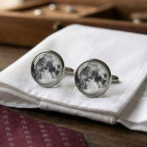 May include: A pair of silver cufflinks with a detailed image of the moon's surface. The cufflinks are round with a glass-like covering, resting on a white folded cloth. A dark red patterned tie is visible in the foreground, with a wooden box in the background.