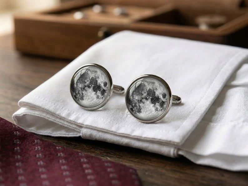 May include: A pair of silver cufflinks with a detailed image of the moon's surface. The cufflinks are round with a glass-like covering, resting on a white folded cloth. A dark red patterned tie is visible in the foreground, with a wooden box in the background.