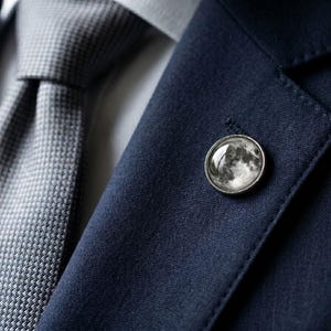 May include: A close-up of a navy blue suit jacket with a gray tie. A silver lapel pin with a detailed moon design is affixed to the jacket. The tie has a textured pattern.